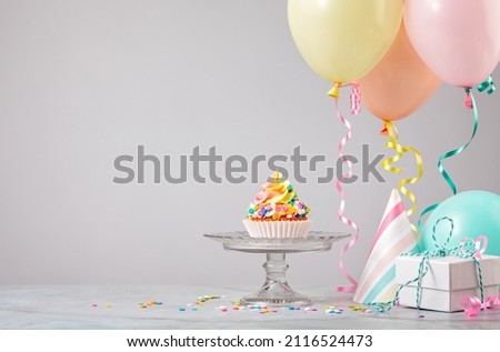 Rainbow Birthday cupcake on a stand with presents, hats and colorful balloons over light grey background. Scene from a birthday party! Royalty-Free Stock Photo #2116524473
