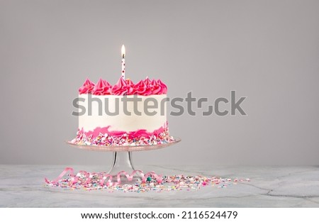 Birthday Cake with pink and white buttercream icing, colorful sprinkles and lit birthday candle over a light grey background.