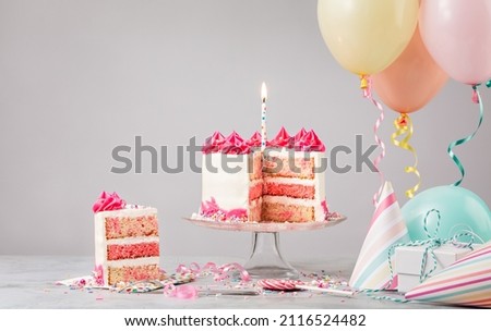 Sliced pink funfetti Birthday cake with candle, presents, hats and colorful balloons over light grey background. Scene from a birthday party!