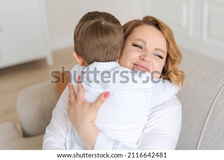 Happy cute caucasian little son hugs mom while sitting on sofa in living room at home. A thousand-year-old mother enjoys the rest with her child, hugs the child. Motherhood concept