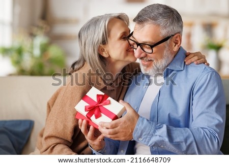 Loving senior woman wife giving  present gift box  for Valentine's day   to happy surprised husband, sitting together on couch at home, smiling mature  man receiving  present. Happy Valentine's day. Royalty-Free Stock Photo #2116648709