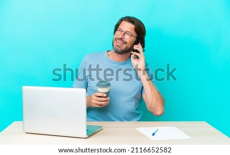 Senior dutch man in a table with a laptop isolated on blue background holding coffee to take away and a mobile