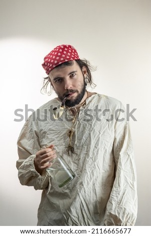 Portrait of a young dark-haired man with a beard, wearing a red cloth bandana on his head and an old shirt with a bottle of rum and a smoking pipe in his teeth on a light background. Pirate Costume,