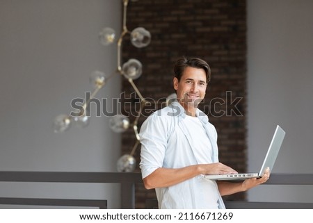Young smiling man looking at camera with laptop in modern home. Remote work.