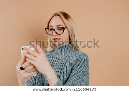 Nice-looking girl with blonde hair is looking at her photo gallery. The girl is standing against the light background