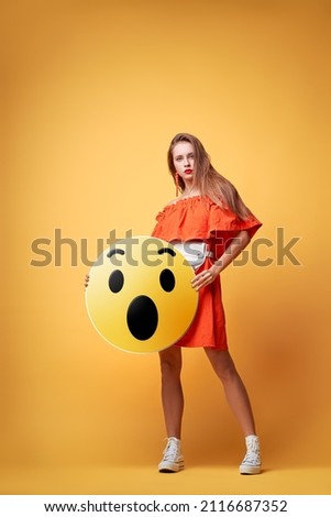 Social network concept. Pretty young woman holding yellow amazed emoji face. Colorful studio portrait.