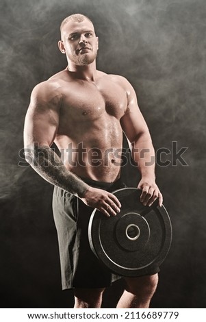 Sports concept, weightlifting, bodybuilding. Portrait of a muscular handsome athletic man holding a large iron disk in his hands. Black background with smoke.