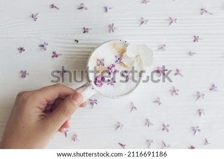 Hand holding delicious coffee with lilac petals flat lay on white wooden background. Happy mothers day. Good morning. Beautiful  flowers in stylish cup of coffee.