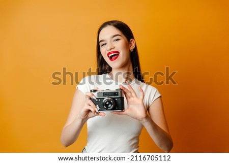 Beautiful brunette is standing against the bright background with a camera in her hands. The girl has black hair and red lips