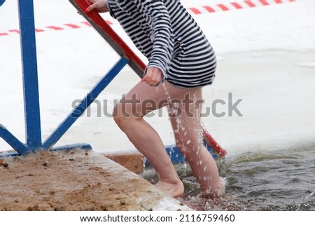 Wet slender white young woman in a striped vest comes out on Pier of the icy water from ice hole. Winter sports outdoor.