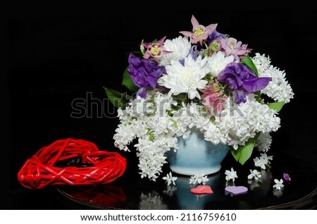 A bouquet of spring flowers with white lilac in a ceramic pot and a red braided heart on a black background with reflections. Valentine's Day Concept