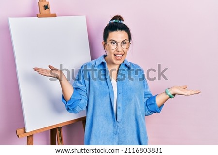 Young hispanic woman standing by painter easel stand smiling showing both hands open palms, presenting and advertising comparison and balance 