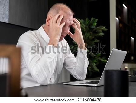 Tired or upset mature businessman sitting at desk and working on laptop. Employee thinking of deadline or workload or suffering from headache. Work overload concept. High quality photo Royalty-Free Stock Photo #2116804478