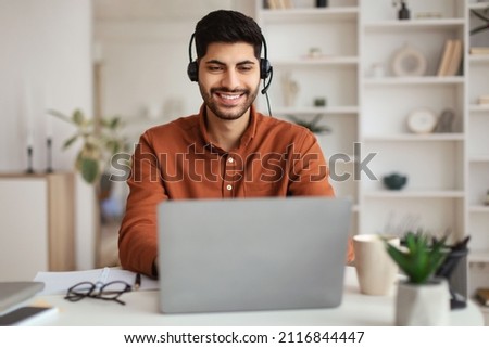 Portrait Of Smiling Middle Eastern Male Manager In Headset Working On Laptop Computer In Modern Home Office, Happy Guy Sitting At Desk And Consulting Clients Online Using Pc, Free Copy Space Royalty-Free Stock Photo #2116844447