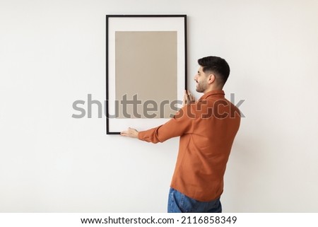 Modern Home Interior And Domestic Decor. Smiling young man hanging painting, putting photo picture frame on the wall. Casual guy holding showing empty poster, design mockup, blank free copy space