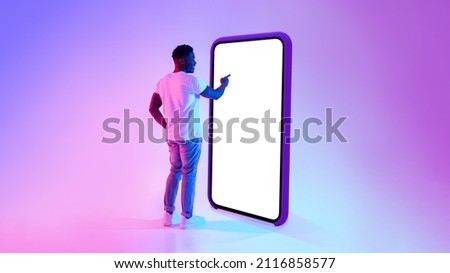 Full length of millennial black guy standing near big cellphone with mockup for your app on screen, interacting with user interface in neon light. Smartphone display template for website or ad Royalty-Free Stock Photo #2116858577