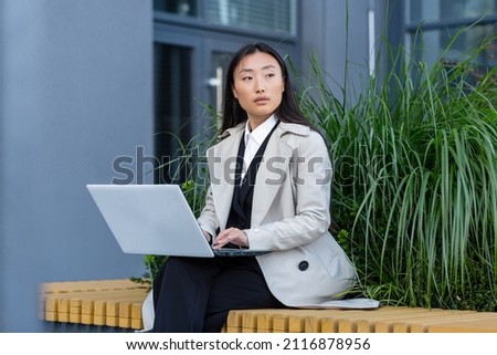 Asian business woman working with laptop outside office, sitting on bench during work break