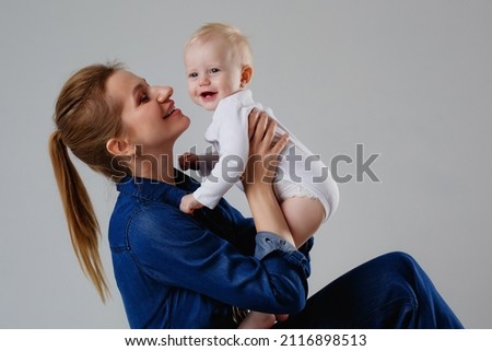 A woman in a denim jumpsuit holds a child in a white bodysuit. Mother and son in the studio on a gray background. Motherhood concept. Unconditional love.