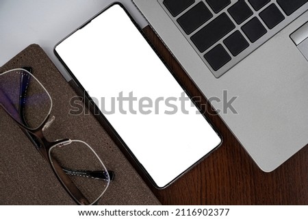 Top view of phone on table near laptop, notepad and glasses with white screen. 