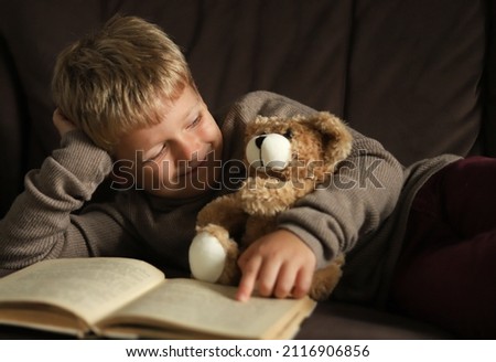 The boy is lying on the sofa reading a book and hugging a teddy bear . Royalty-Free Stock Photo #2116906856