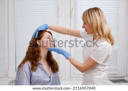 A young red-haired beautiful girl undergoes a medical examination in a cosmetology clinic. A female dermatologist consultation. Close up. Royalty-Free Stock Photo #2116907840