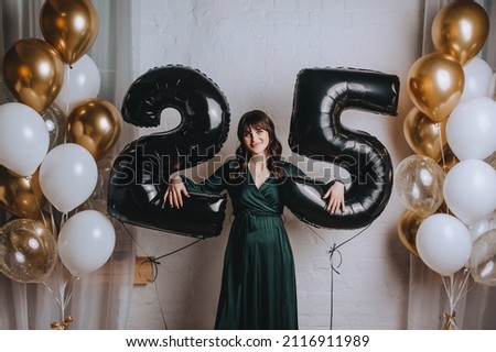 A beautiful, smiling brunette girl in a green dress holds the number 25 from balloons for her birthday.