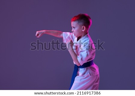 Pupil. Portrait of kid, male taekwondo, karate athletes in doboks doing basic movements isolated on purple background in neon. Concept of sport, education, skills, martial arts, healthy lifestyle. Royalty-Free Stock Photo #2116917386