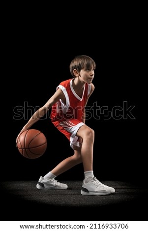Portrait of young boy in red uniform training, playing basketball isolated over black background. Popular game. Concept of sport, active lifestyle, health, team game, youth and ad