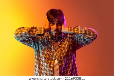 Portrait of young boy covering ears with hands isolated over gradient yellow orange background in neon. Concept of youth, emotion, facial expression, fashion, lifestyle, ad