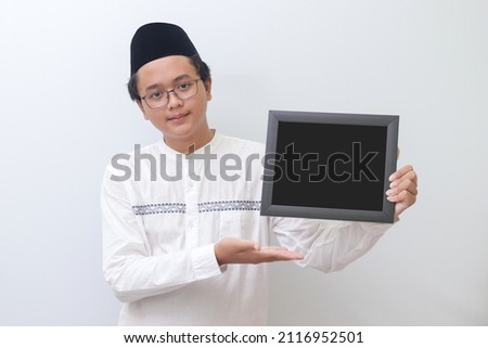 Portrait of young Asian muslim man holding a blackboard with hand pointed. Blank space for typography or text to fill. Isolated image on white background