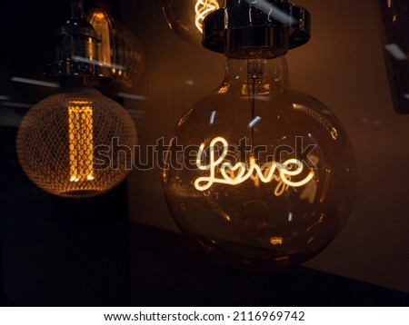Light bulb with the word LOVE in it. A glass sphere bulb on display in a store. Another orange bulb behind it, with dark background. The word is written in fancy calligraphy font. Concept for love.