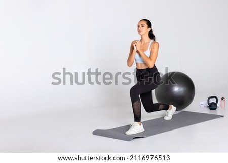 female trainer shows exercise lunge with hands in front of her chest on grey mat and with gym equipment in sport wear on white background, woman workout with knee bent for stretching