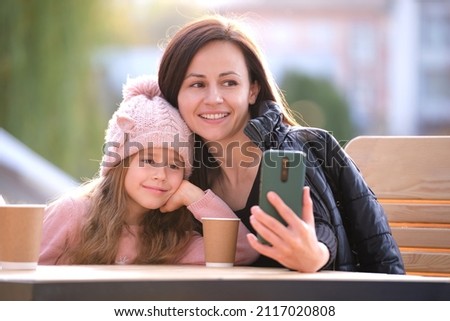 Young mother and her child daughter taking picture with phone selfie camera sitting at street cafe with hot drinks on sunny fall day. Social media presence in everyday life concept
