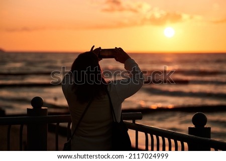 Female shooting sunset with smartphone, woman taking photos of beautiful sunset with mobile phone. Young woman taking pictures of evening seascape and bright sunset, mobile photography
