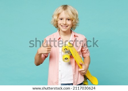 Smiling little curly kid boy 10s years old wearing pink shirt hold yellow skateboard showing thumb up isolated on blue turquoise color background children studio portrait. Childhood lifestyle concept