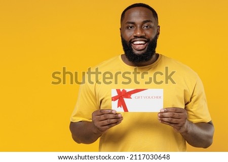 Young smiling happy black man 20s wearing bright casual t-shirt hold gift certificate coupon voucher card for store isolated on plain yellow color background studio portrait. People lifestyle concept