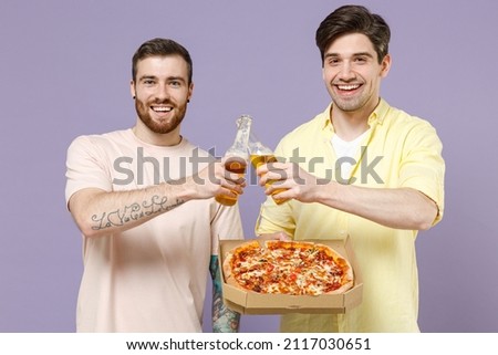 Two young men friends together in casual t-shirt eat italian pizza in cardboard flatbox drink beer clink isolated on purple background studio portrait People lifestyle concept. Tattoo translate fun