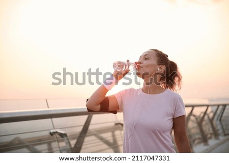 Happy determined athlete, beautiful Hispanic woman wearing pink t-shirt, smartphone holder and terry wristbands stands on a city bridge and drinks water, resting after early morning jog at sunrise Royalty-Free Stock Photo #2117047331