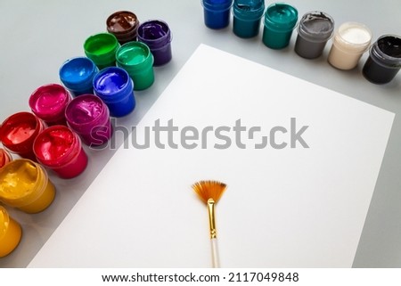 A set of open jars with gouache in different colors and a brush on a white sheet of paper. Hobby - drawing. High quality photo