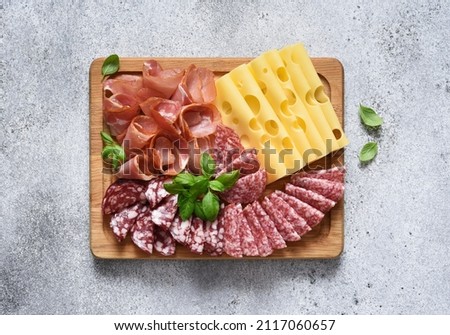 Delicacies. Cheese, prosciutto, salami on a wooden square board on a concrete background. Royalty-Free Stock Photo #2117060657