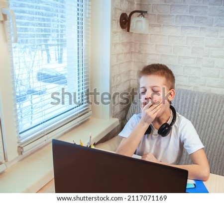 boy yawning while using a laptop in a kitchen. Distance learning online.