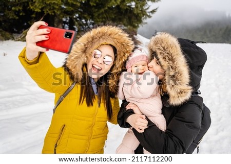 Two caucasian women and baby girl taking selfies mother with her small child and her friend or aunt taking photos or making a video call using mobile phone in winter day in nature with snow outdoor