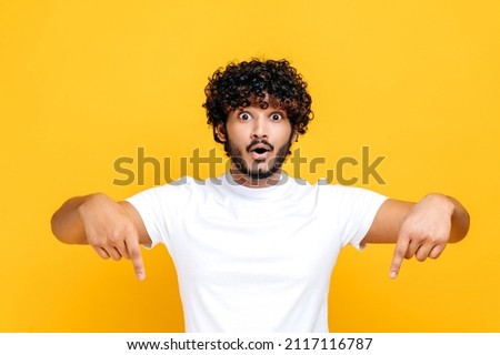 Positive indian or arabian happy guy in basic white t-shirt, amazed looks at camera and points fingers down, surprised face expression, stands on isolated orange color background Royalty-Free Stock Photo #2117116787