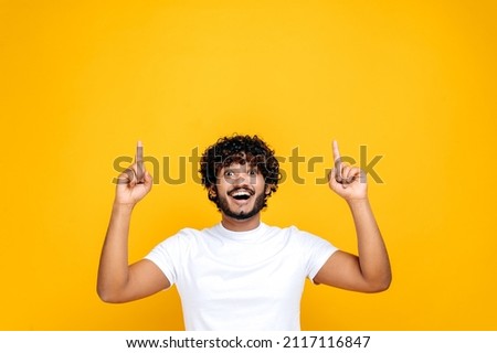 Happy indian or arabian guy in basic white t-shirt, amazed looks at the camera and points fingers up, at empty space, stands on isolated orange color background. Mock-up concept Royalty-Free Stock Photo #2117116847