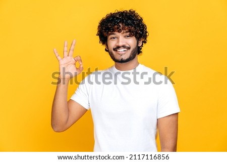 Happy pleasant indian or arabian curly-haired guy in white t-shirt, showing ok sign while standing on isolated orange background, looks at camera, smiling friendly