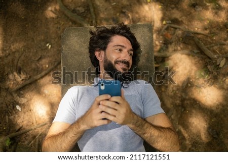 View of young man using a smartphone at day time lying down on a bench at a park. High quality photo. Mobile phone, technology, urban, nature  concept. High quality photo