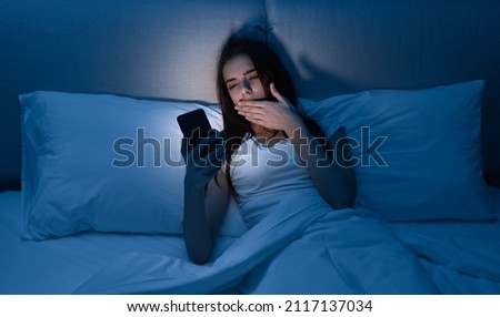 Tired woman covering mouth and yawning while lying in bed and browsing social media on mobile phone at night at home Royalty-Free Stock Photo #2117137034