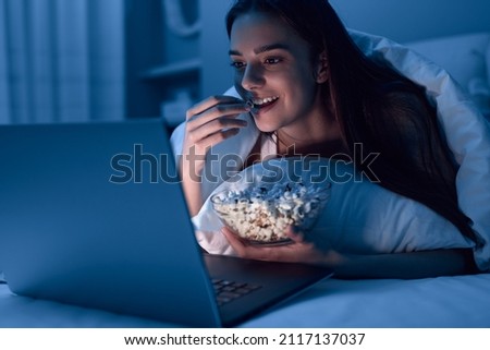 Happy young woman eating popcorn while lying on bed under blanket and watching movie on netbook at night at home Royalty-Free Stock Photo #2117137037