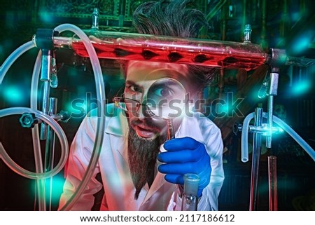 Portrait of a funny mad scientist conducting scientific experiments in the laboratory and looking at the camera through a magnifying glass. Fantasy, science fiction novel. Royalty-Free Stock Photo #2117186612
