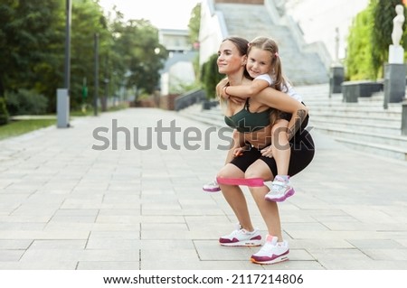 Athletic mom exercising with her little daughter on her back outdoors. Squat with fitness rubber bands. Healthy lifestyle, fitness active family concept. Training together Royalty-Free Stock Photo #2117214806